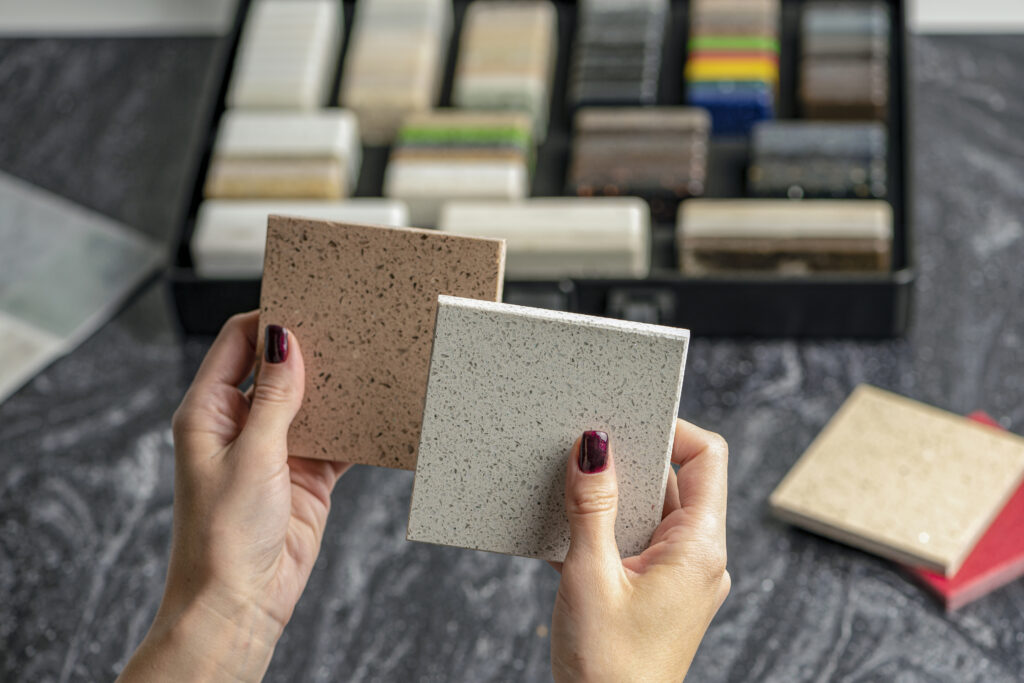 Deux mains tiennent des échantillons de matériaux de couleur beige et grise, avec d'autres échantillons colorés en arrière-plan.