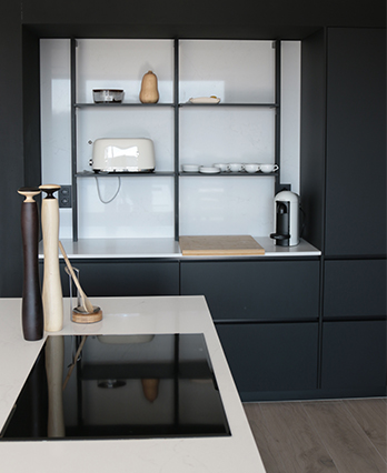 Cuisine moderne avec placards noirs, étagères ouvertes, grille-pain blanc, cafetière, plan de travail en bois clair et décorations en bois sur l’îlot central.