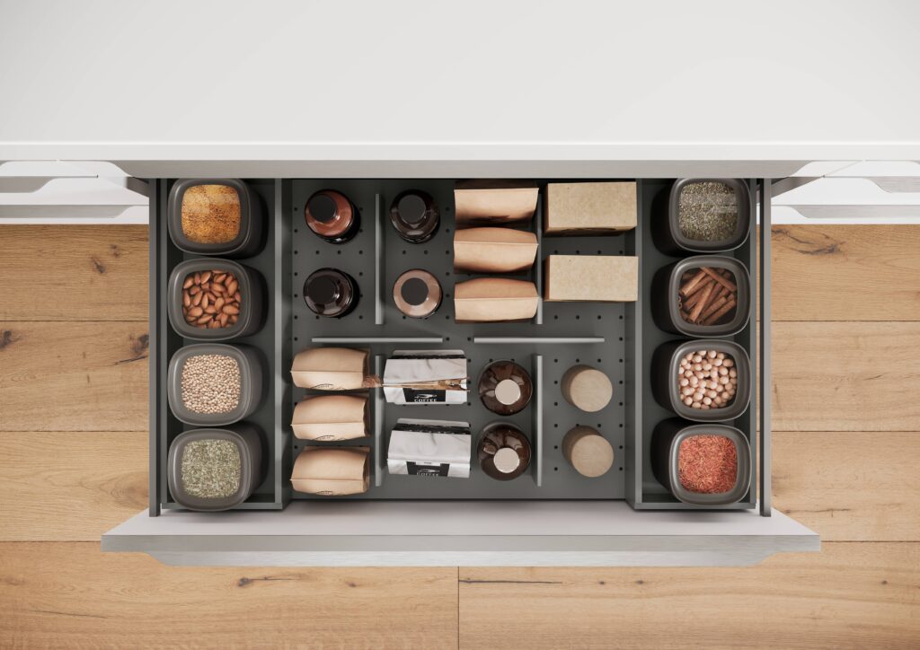 Tiroir de cuisine ouvert avec des bocaux gris contenant des épices et des graines, et des paquets en papier brun alignés.