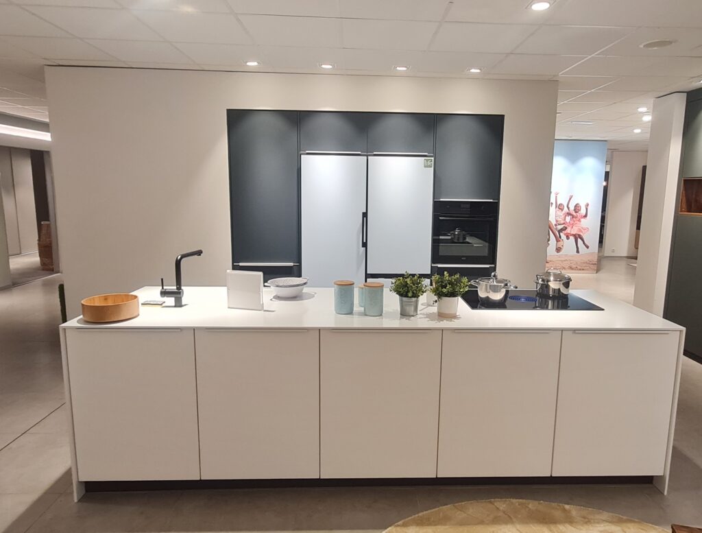Cuisine moderne avec îlot central blanc, électroménager encastré noir, pots verts, et casseroles en acier inoxydable sur plaque de cuisson.