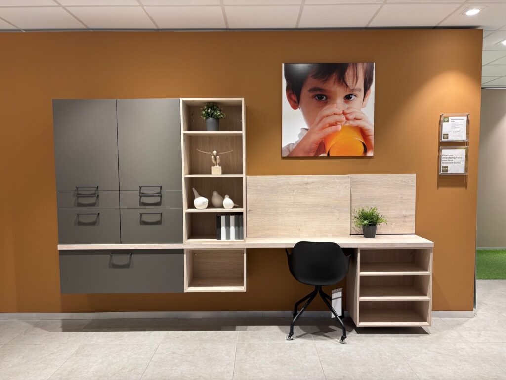 Meuble mural gris avec étagères décorées, bureau en bois clair avec chaise noire, et photo d'enfant buvant dans un verre orange.