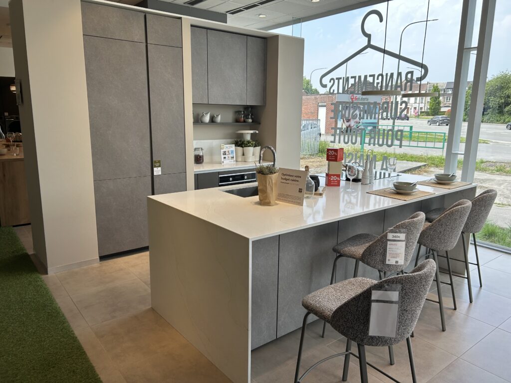 Une cuisine moderne avec îlot central blanc, chaises grises, placards gris et fenêtres vitrées donnant sur l'extérieur.