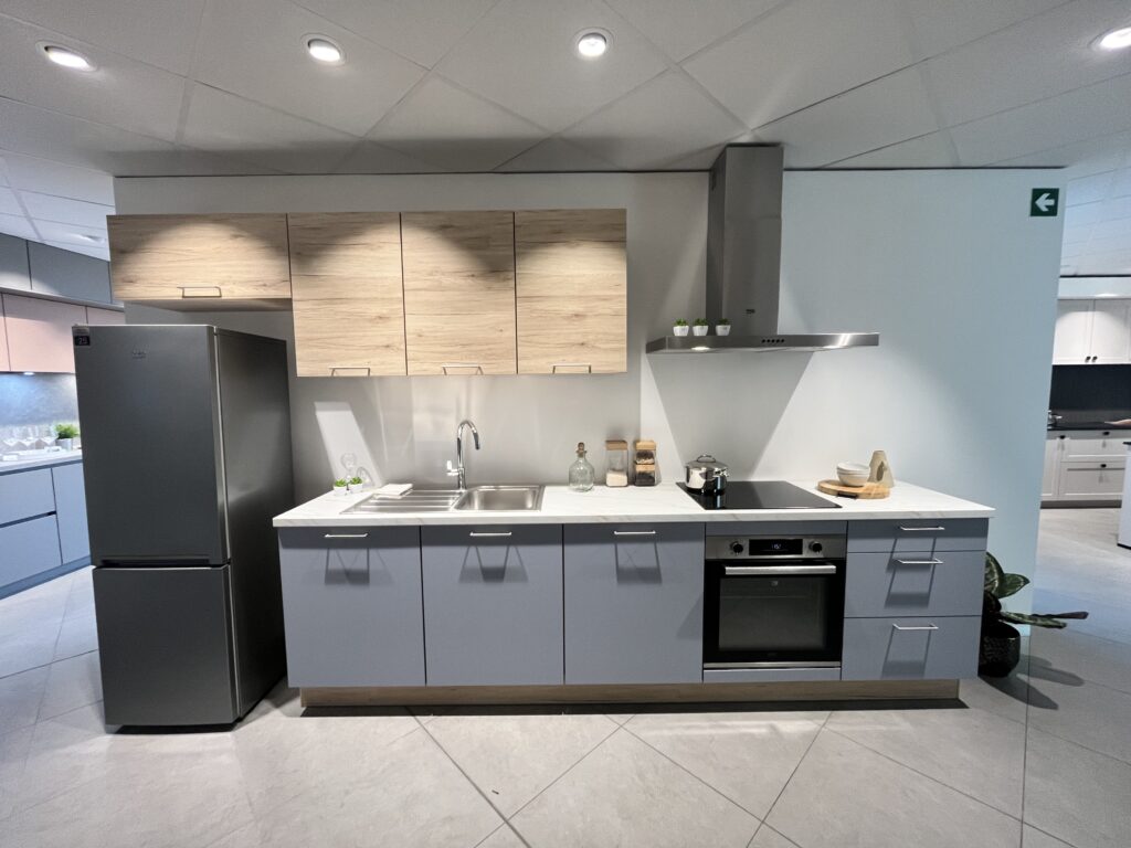Cuisine avec meubles bas aux façades de couleur bleue et meubles hauts aux façades en bois. Frigo posé, four encastré dans meuble bas et hotte apparente.