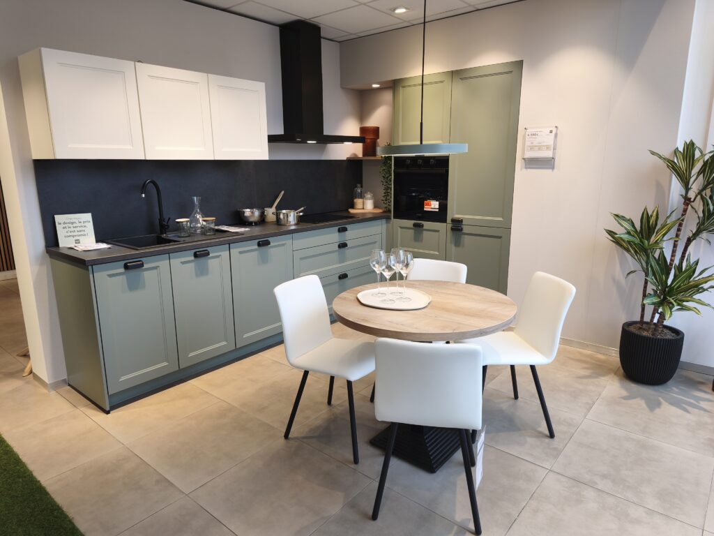 Cuisine cottage avec façades de couleur vert/bleu. Meubles hauts aux façades blanches. Crédence noire. Coin repas avec table ronde en bois et 4 chaises blanches.