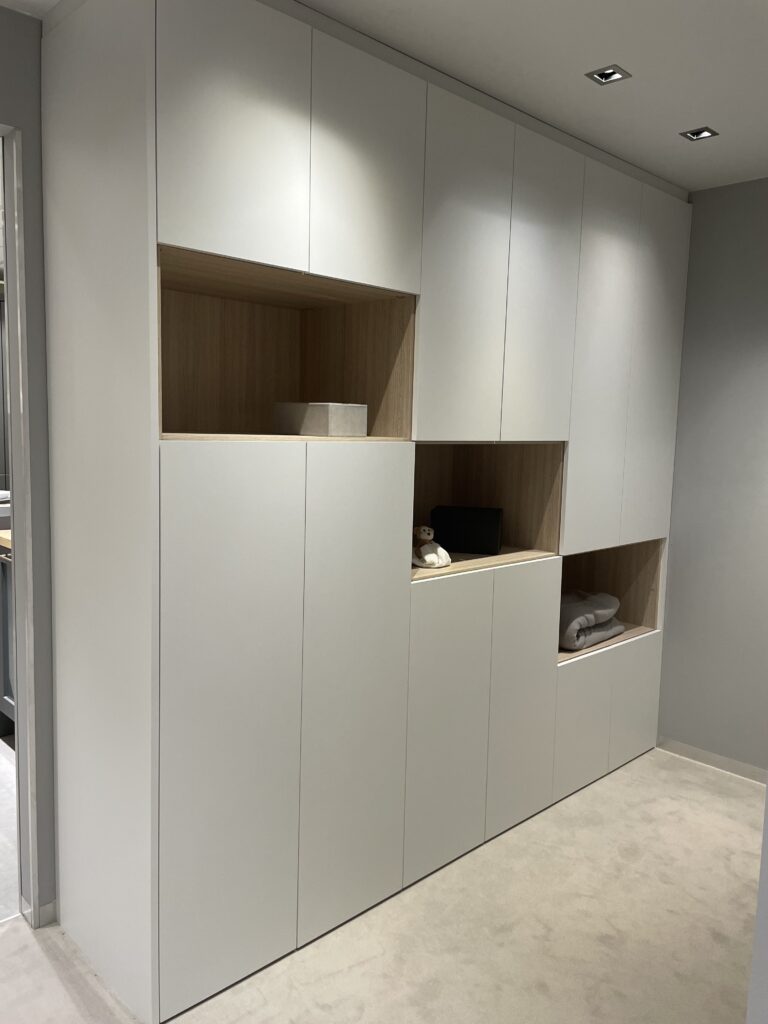 Grand meuble sur mesure blanc avec niches ouvertes en bois clair, disposé le long d’un mur gris, avec objets décoratifs et linge plié.