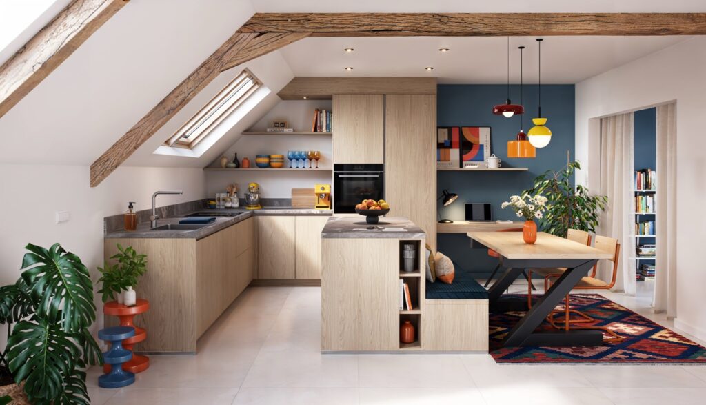 Cuisine sous combles en bois clair avec plan de travail gris, îlot central, table intégrée, banquette bleue, poutres apparentes et suspensions colorées.