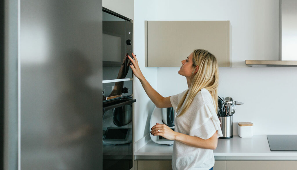 Une femme en t-shirt blanc règle un four encastré noir dans une cuisine aux meubles beiges et plan de travail gris.