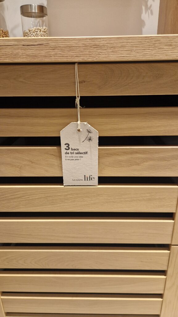 Meuble de cuisine en bois clair à lattes horizontales avec une étiquette blanche suspendue au centre. Deux bocaux transparents sont posés sur le plan de travail en haut à gauche.