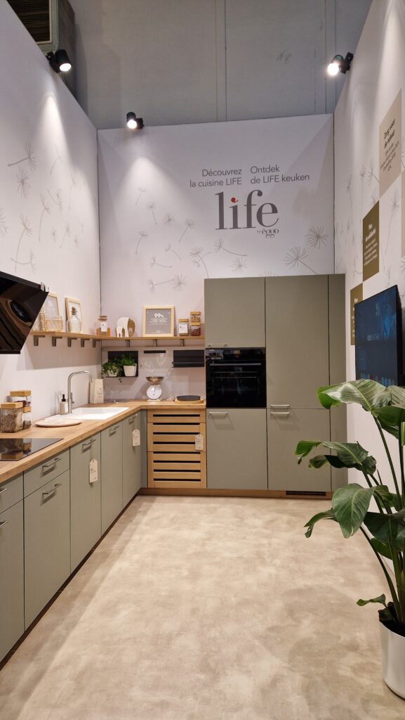 Cuisine en L avec meubles vert sauge, plan de travail en bois clair, évier blanc, colonnes avec four noir et plante verte au premier plan.