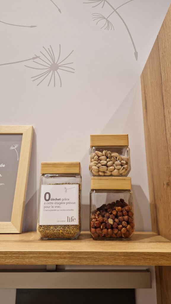 Trois bocaux en verre avec couvercle en bois, remplis de lentilles, pistaches et noisettes, sont posés sur une étagère en bois contre un mur clair décoré.