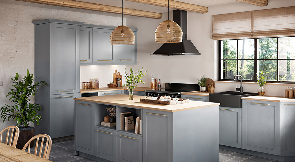 Cuisine équipée avec meubles gris bleu, plan de travail en bois clair et îlot central. Une hotte noire, un évier noir sous la fenêtre et deux suspensions en osier sont visibles.