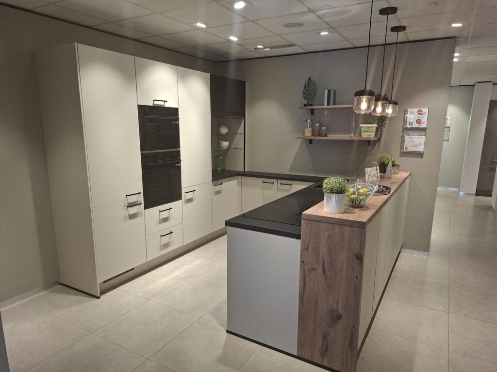 Cuisine moderne en L avec armoires blanches, plan de travail noir et îlot central avec extrémité en bois clair. Des étagères murales et trois suspensions noires complètent l’ensemble.