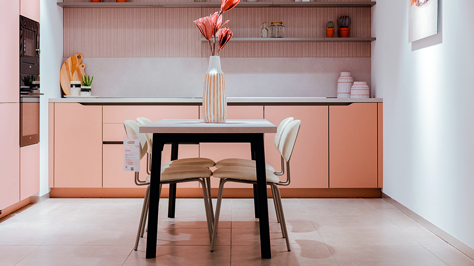 Cuisine moderne aux meubles rose pâle avec table blanche au centre, entourée de quatre chaises claires. Une grande vase blanche avec fleurs roses est posée sur la table.