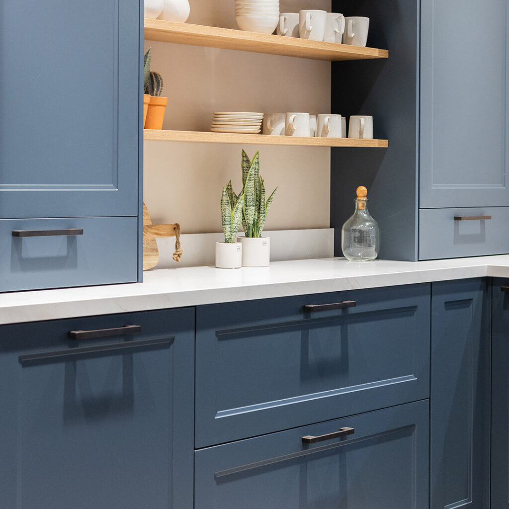 Cuisine avec placards bleus et plan de travail blanc, encadrant des étagères en bois clair avec assiettes, tasses blanches, deux plantes en pot et une bouteille en verre.