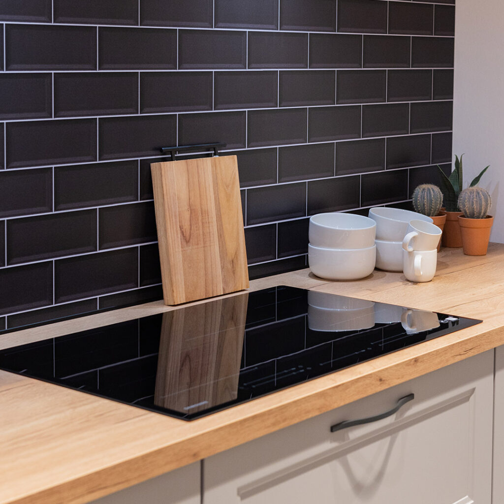 Plan de travail en bois clair avec plaque de cuisson noire, devant une crédence en carreaux noirs. Une planche, des bols blancs, des tasses blanches et deux cactus y sont posés.