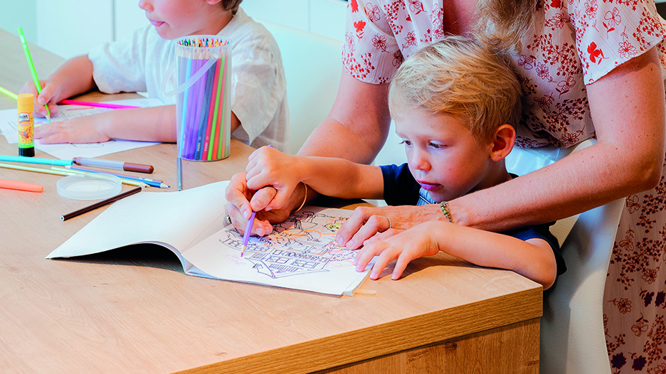 Un enfant blond colorie un dessin sur un plan de travail en bois clair, accompagné d’une femme. Des feutres et crayons de couleur sont posés à gauche.