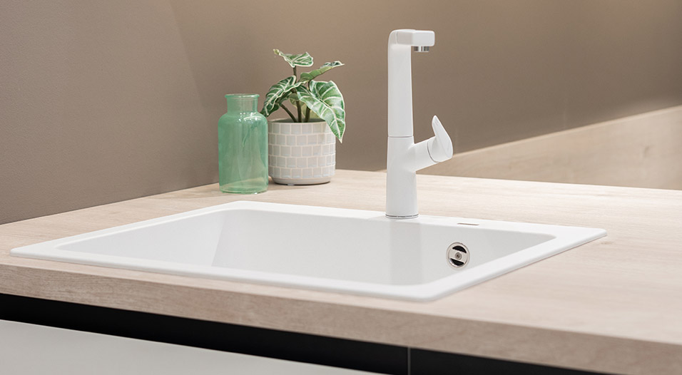 Évier encastré blanc avec robinet blanc sur un plan de travail en bois clair, accompagné d’un vase vert translucide et d’une plante en pot.