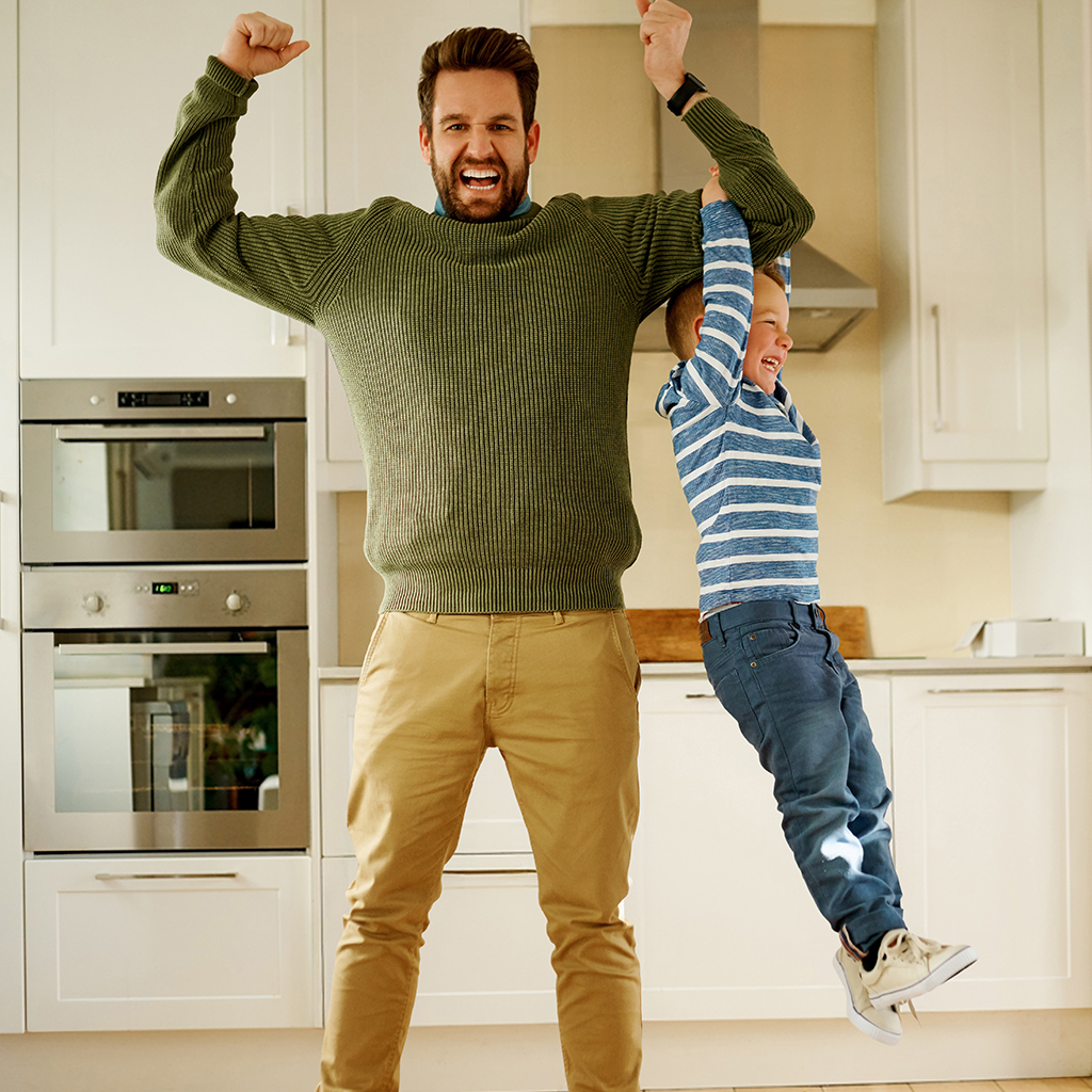Un homme en pull vert et pantalon beige lève les bras dans une cuisine blanche, à côté d’un enfant en haut rayé bleu et blanc.