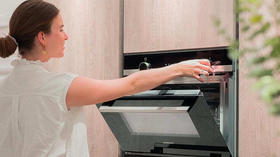 Une femme en blouse blanche ouvre un four encastré noir et inox Siemens, intégré en hauteur dans une colonne de cuisine en bois clair.