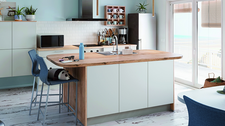 Cuisine moderne avec îlot central blanc et plan de travail en bois, deux chaises bleues, électroménagers noirs et baie vitrée à droite.
