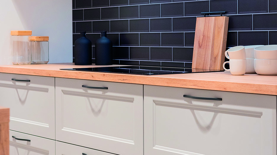 Plan de travail en bois clair sur meubles blancs avec poignées noires, plaque de cuisson noire, pots en verre, bouteilles noires, planche en bois et tasses blanches.