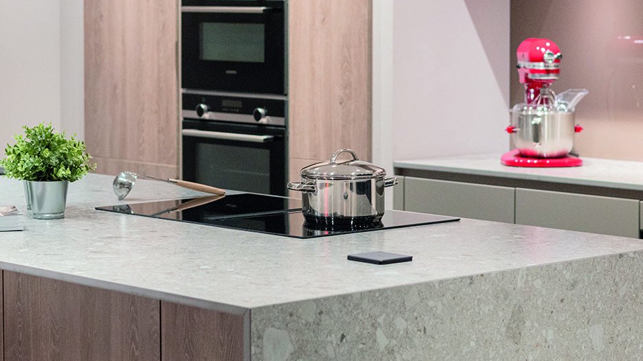Îlot de cuisine en pierre claire avec plaque de cuisson noire et casserole en inox au centre, devant des colonnes en bois clair avec four noir encastré.