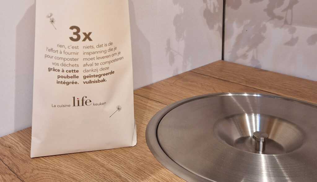 Poubelle intégrée ronde en métal avec couvercle sur un plan de travail en bois, à côté d’un sac en papier blanc posé à gauche.