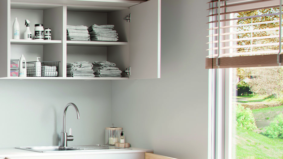 Buanderie avec meuble mural blanc ouvert contenant des serviettes pliées et produits ménagers, au-dessus d’un évier blanc avec robinet chromé, à côté d’une fenêtre avec store.