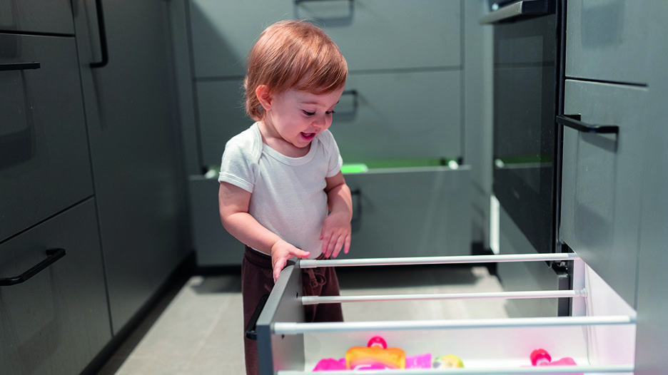 Un enfant en body blanc se tient devant un tiroir ouvert dans un meuble gris foncé, avec des jouets colorés visibles à l’intérieur.