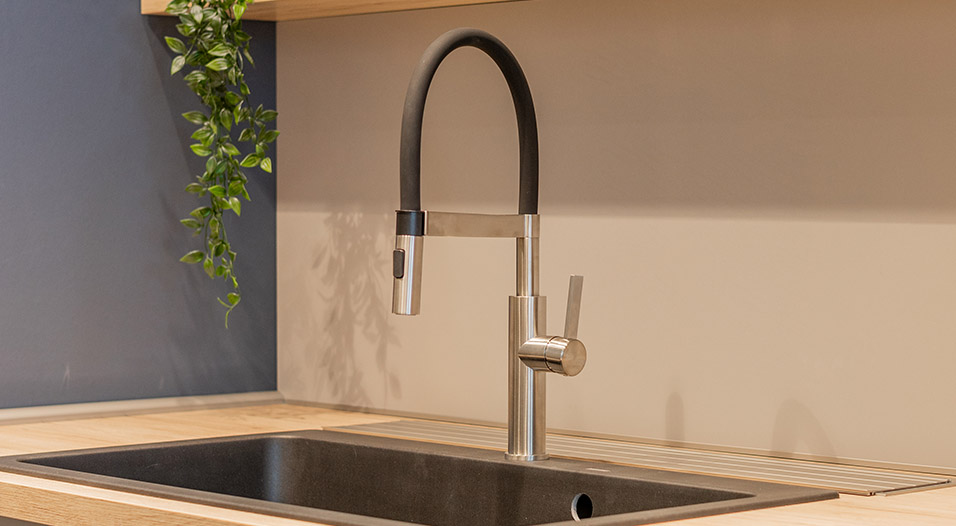 Robinet de cuisine en métal brossé avec bec noir arqué au-dessus d’un évier rectangulaire noir, posé sur un plan de travail en bois clair.