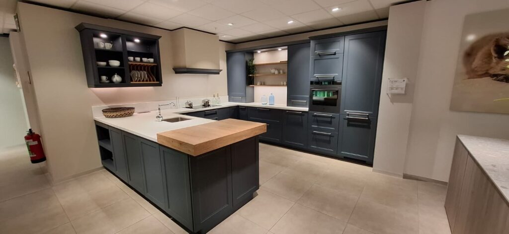Cuisine en U avec meubles bleu foncé, plan de travail blanc, îlot central avec tablette en bois, évier intégré, hotte murale et four encastré.