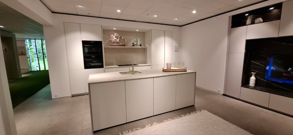 Cuisine moderne blanche avec îlot central et évier intégré, colonnes de rangement et fours noirs à gauche, niche décorative au fond et meuble mural noir à droite.