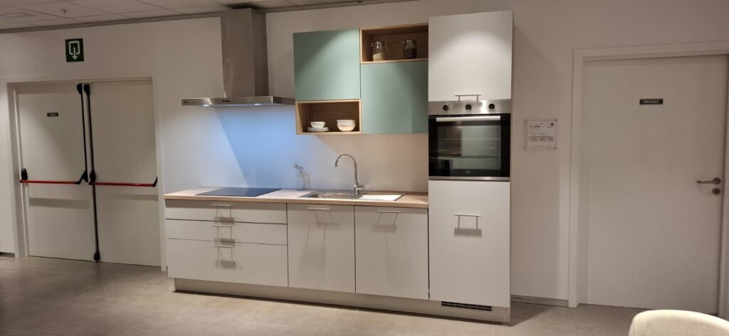 Cuisine équipée linéaire avec meubles blancs, plan de travail en bois clair, placards hauts vert sauge, hotte inox, évier, plaque de cuisson et four encastré à droite.