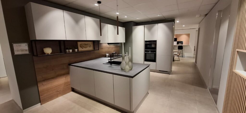 Cuisine moderne avec îlot central blanc et plan de travail gris, meubles hauts blancs, crédence en bois, fours encastrés noirs et trois vases blancs posés au centre.