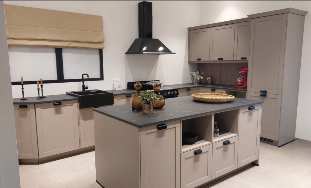 Cuisine équipée beige avec îlot central et plan de travail gris, évier noir sous une fenêtre, hotte noire murale et meubles hauts à droite.