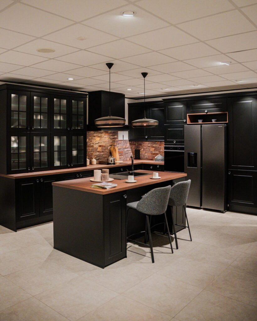 Cuisine noire avec îlot central en bois, deux tabourets gris, armoires vitrées à gauche, réfrigérateur gris à droite et crédence en pierre au fond.