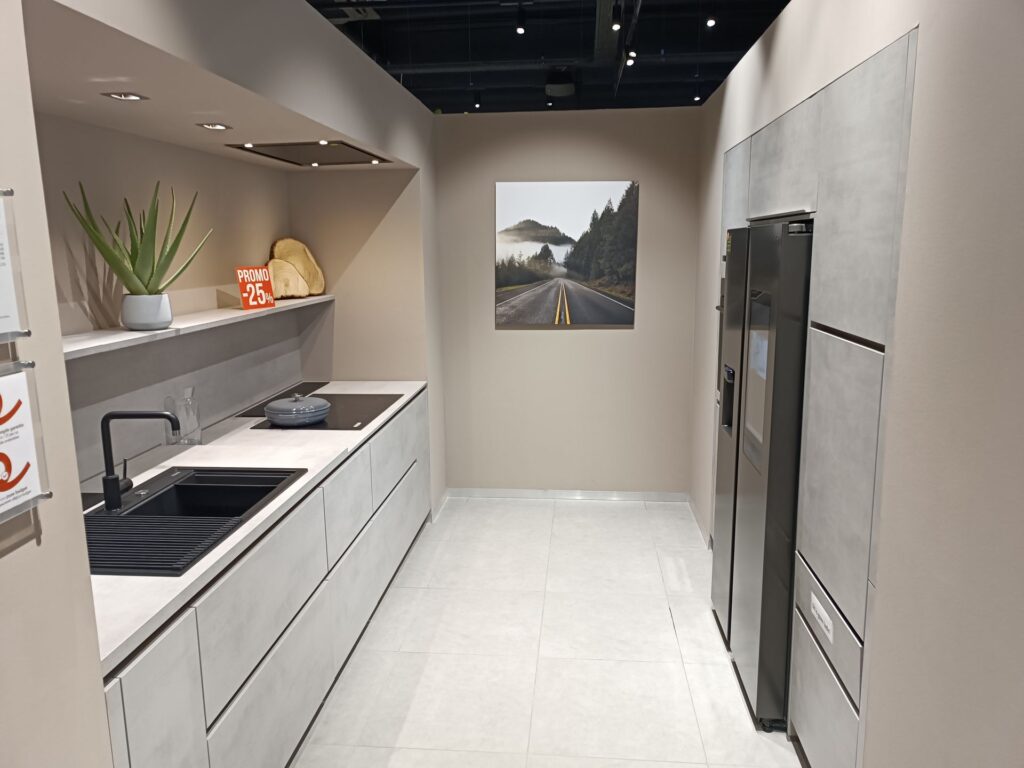 Cuisine étroite aux meubles gris clair avec plan de travail blanc, évier noir et plaque de cuisson à gauche, grand réfrigérateur noir et armoires à droite.