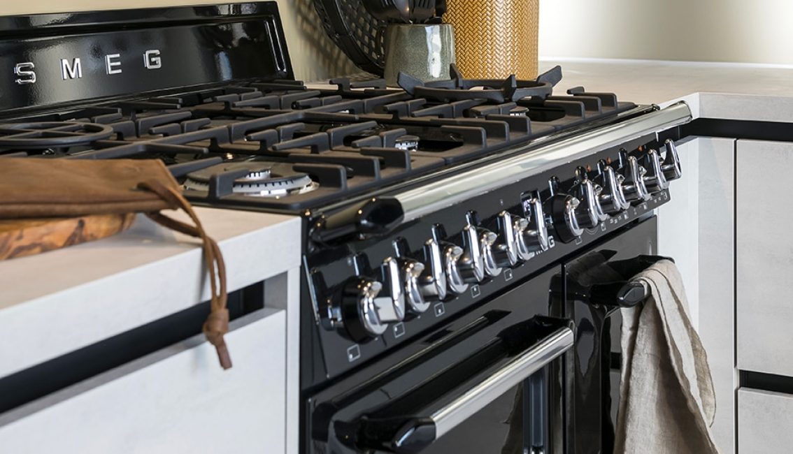 Cuisinière noire avec plusieurs brûleurs et boutons argentés, torchon beige accroché, plan de travail clair avec ustensiles en arrière-plan.