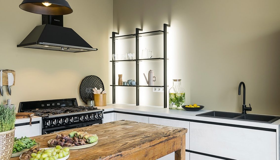 Cuisine moderne avec plan de travail en bois, hotte noire, étagères murales éclairées, et évier noir avec carafe d'eau citronnée.