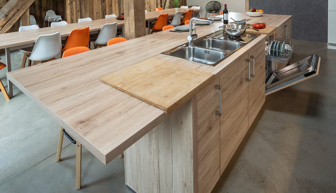 Îlot de cuisine en bois clair avec évier double, planche à découper, lave-vaisselle ouvert et chaises orange et blanches en arrière-plan.
