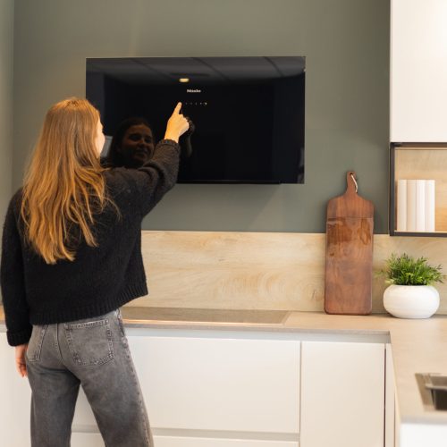 Femme touchant un écran de cuisine Miele noir, avec un plan de travail en bois clair et une petite plante en pot blanc à droite.