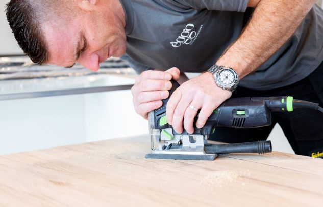 Un homme utilise une scie sauteuse noire et verte sur une planche en bois clair, vêtu d'un t-shirt gris.