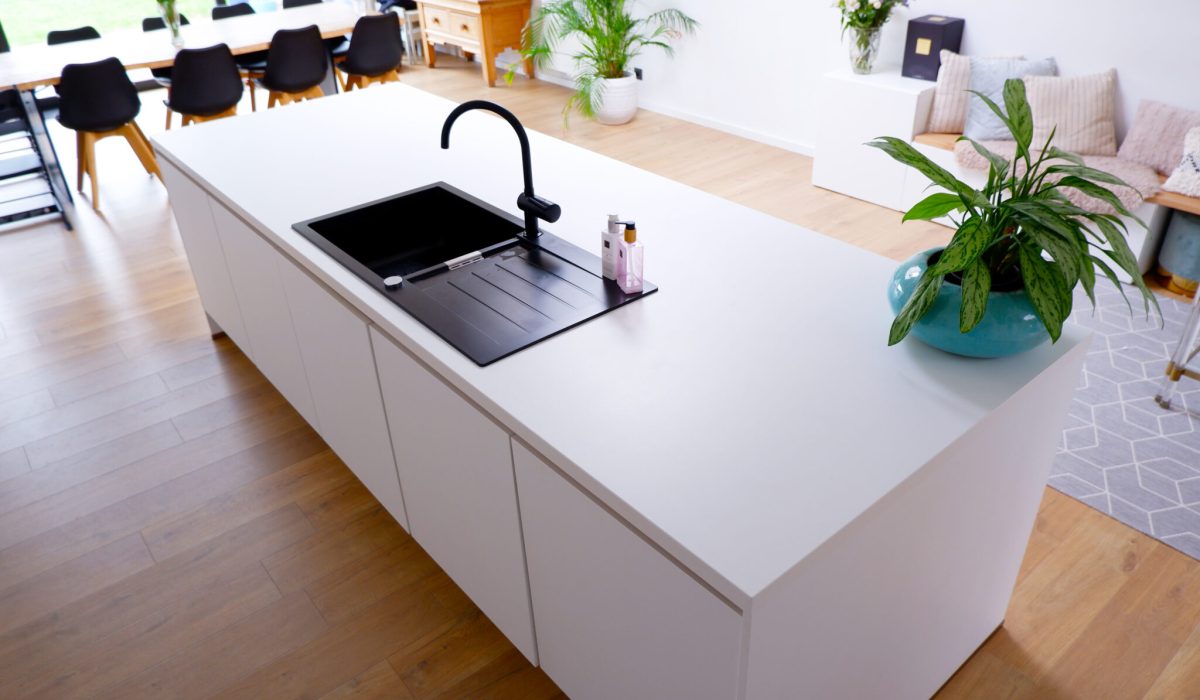 Îlot de cuisine blanc avec évier noir et robinet, plante verte dans un pot bleu, table à manger en arrière-plan.