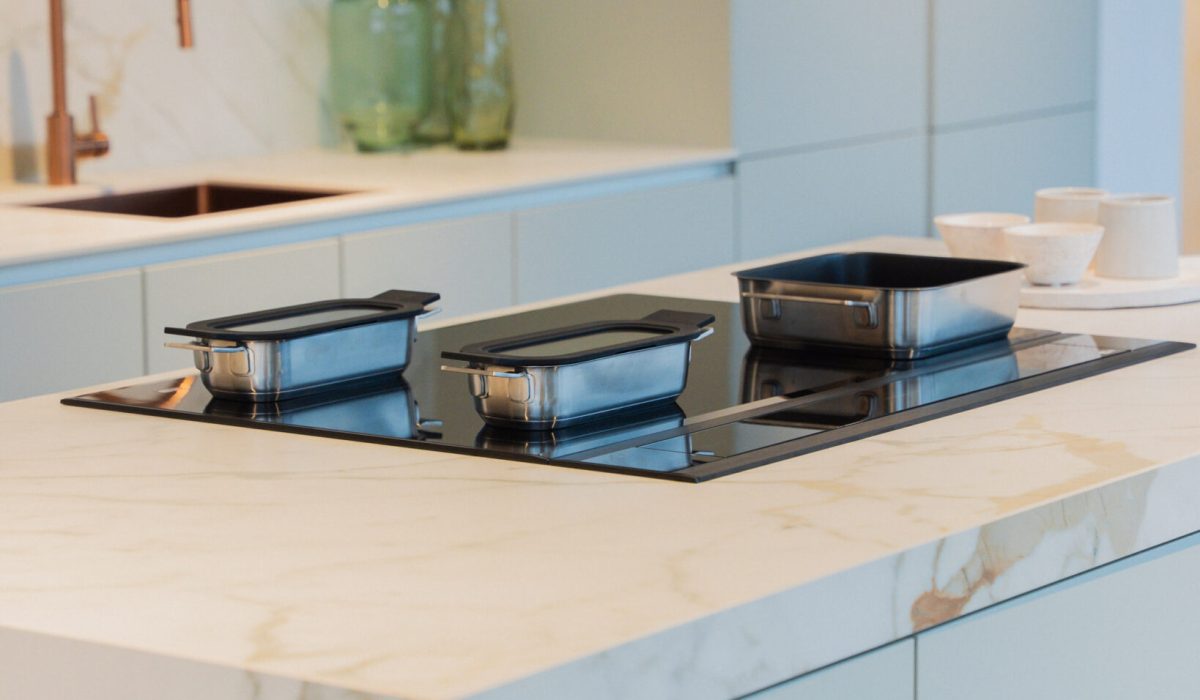 Îlot de cuisine blanc effet marbre avec plaque noire intégrée et trois récipients métalliques, devant un évier noir avec robinet cuivré.