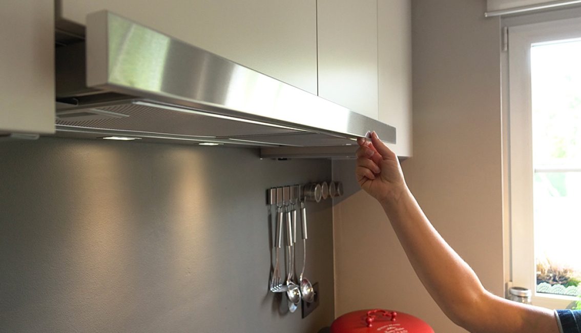 Une main ajuste une hotte de cuisine en métal sous des placards blancs, avec des ustensiles accrochés au mur gris.