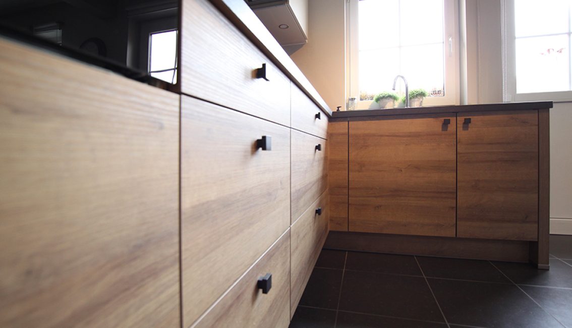 Meubles de cuisine en bois clair avec poignées noires, plan de travail sombre, fenêtre en arrière-plan et plantes vertes près de l'évier.