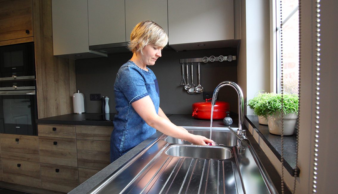Une personne est debout dans une cuisine moderne, rinçant ses mains sous un robinet en acier inoxydable, avec des armoires en bois et des plantes en pot sur le rebord de la fenêtre.