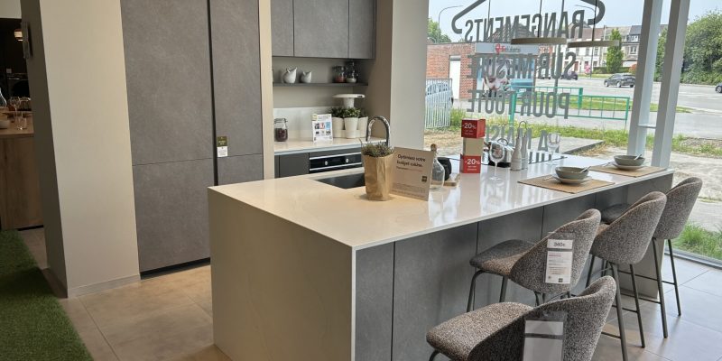 Une cuisine moderne avec îlot central blanc, chaises grises, placards gris et fenêtres vitrées donnant sur l'extérieur.