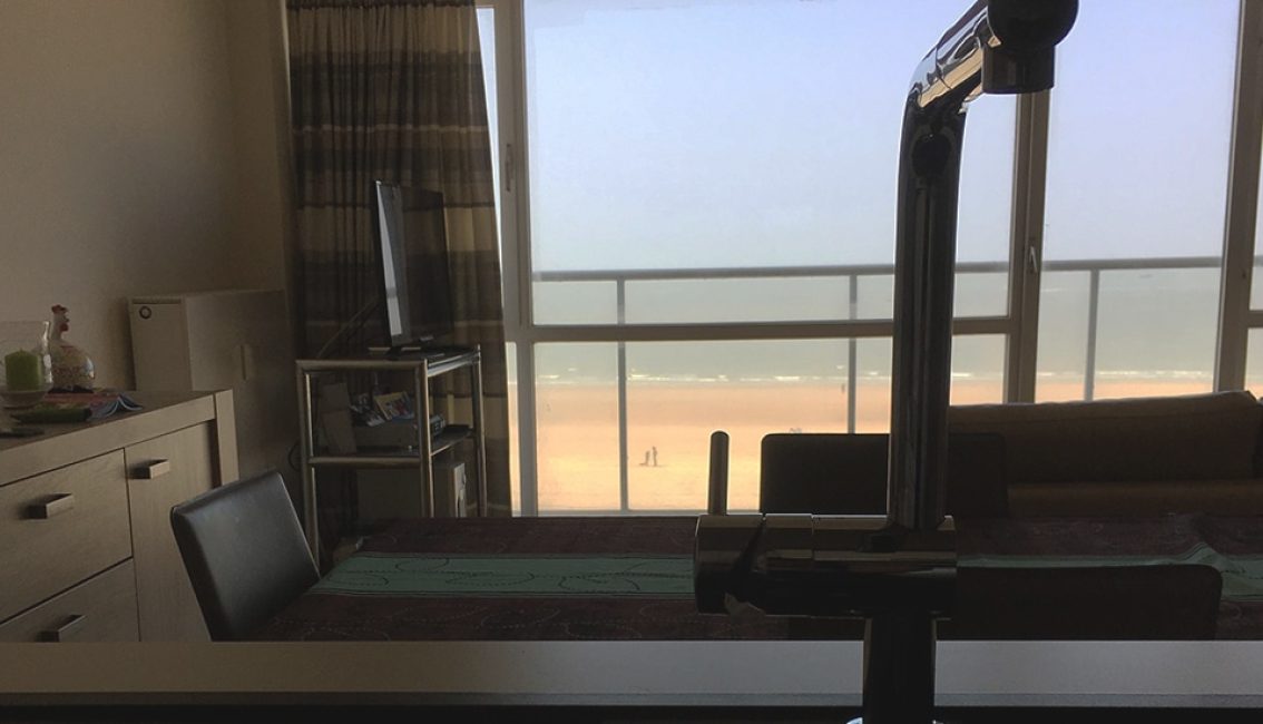 Vue intérieure d'un salon avec table, chaise noire, télévision, et grande fenêtre donnant sur une plage ensoleillée.