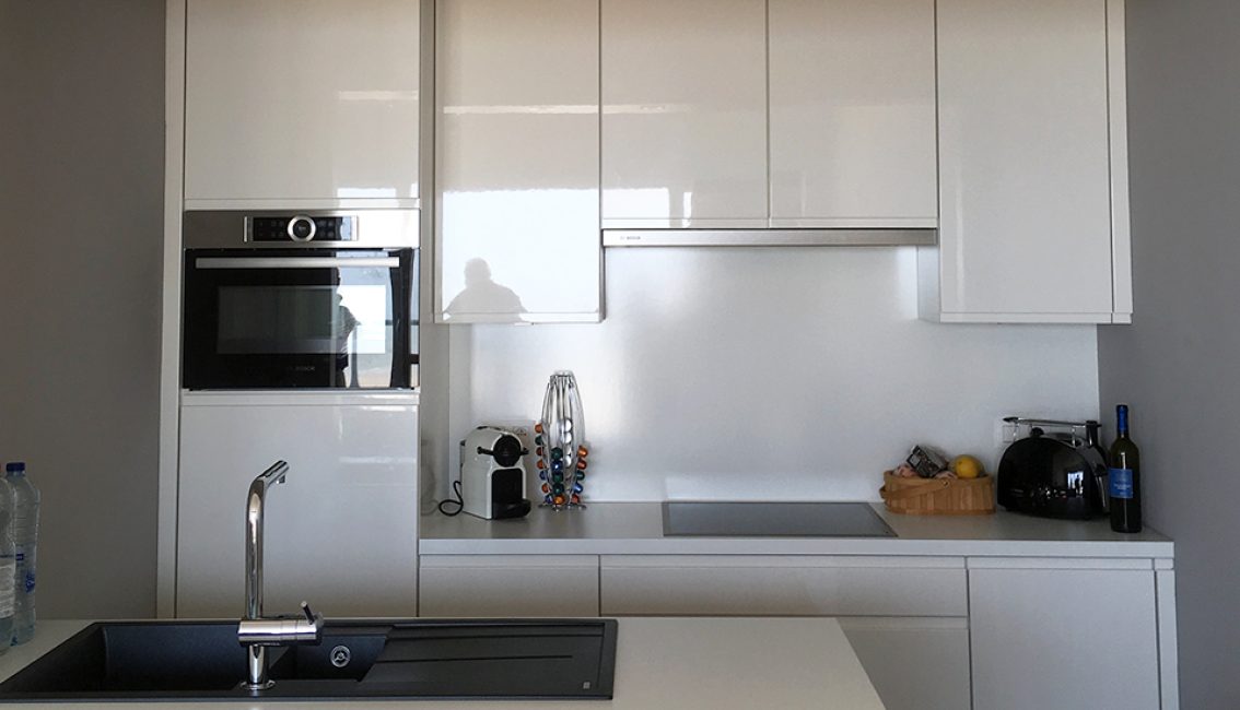 Cuisine moderne avec meubles blancs, four intégré, machine à café, porte-capsules coloré, évier noir, et accessoires sur le plan de travail.